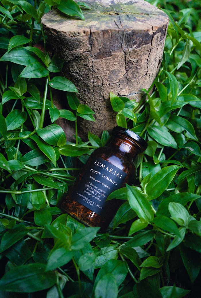 A brown glass bottle with Lumaraki label rests amidst vibrant green foliage and tree stump.