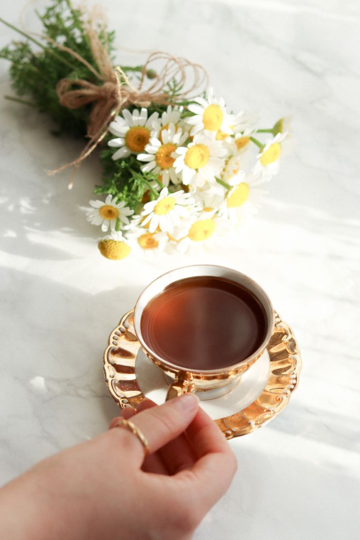 About From above of crop female hand holding ceramic cup with hot tea on table with bouquet of chamomiles near in bright sunlight